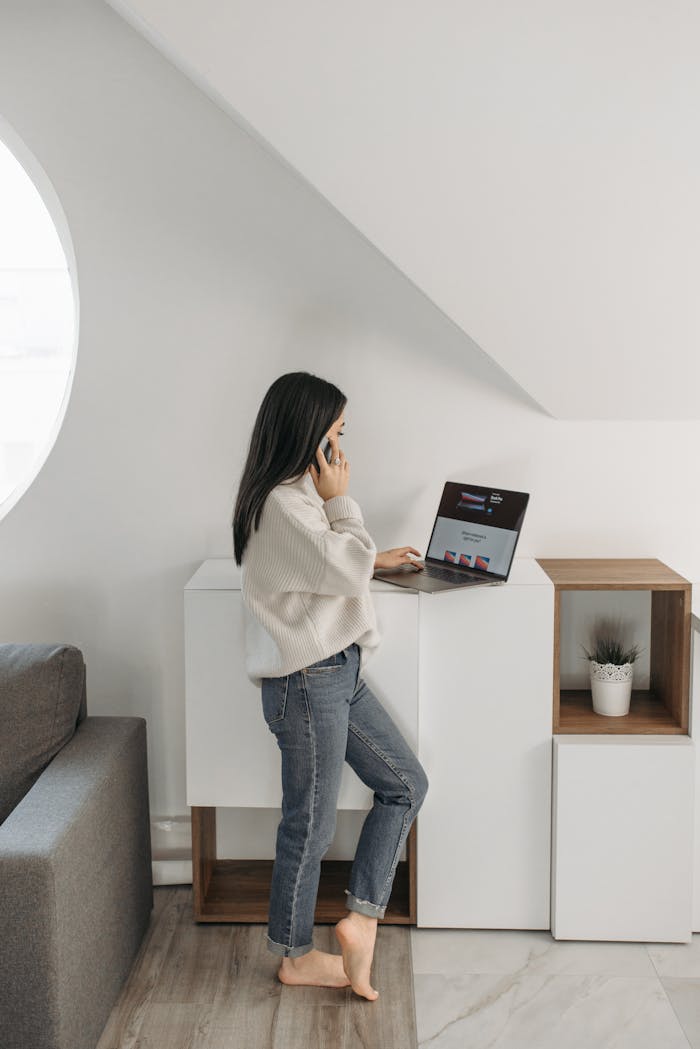 A woman stands indoors wearing a sweater, using a laptop for online shopping.